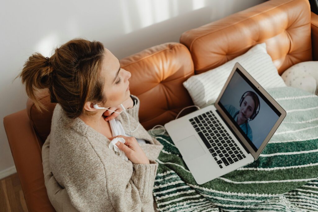 A woman video calling on her laptop, relaxing on a sofa with earphones.