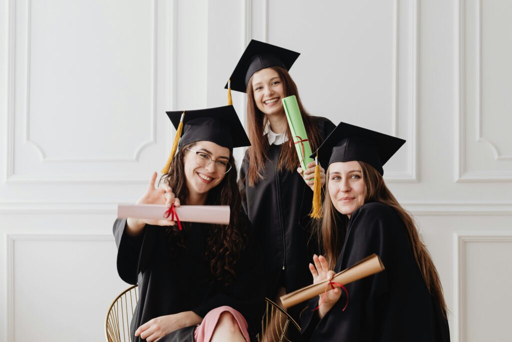 Three young graduates in gowns celebrate with diplomas indoors, exuding happiness.